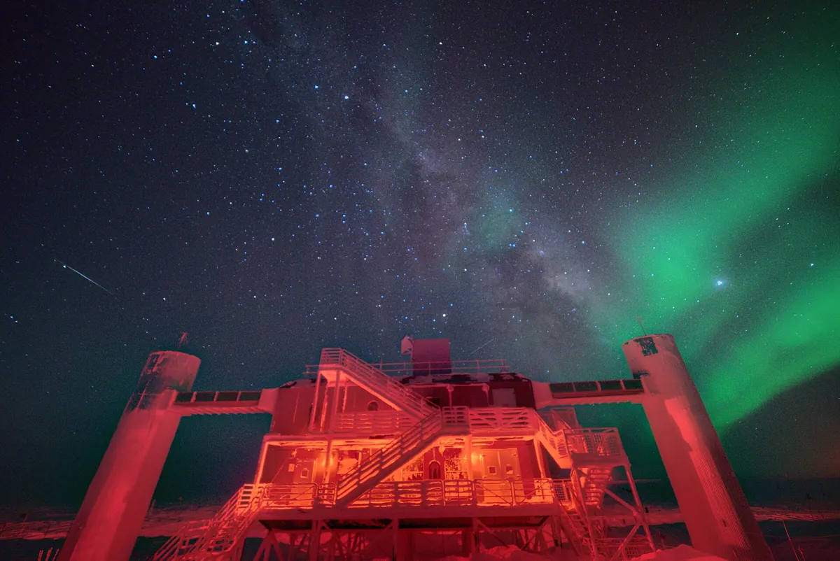 The IceCube Laboratory at the South Pole at night, lit red, under the Milky Way and a green aurora